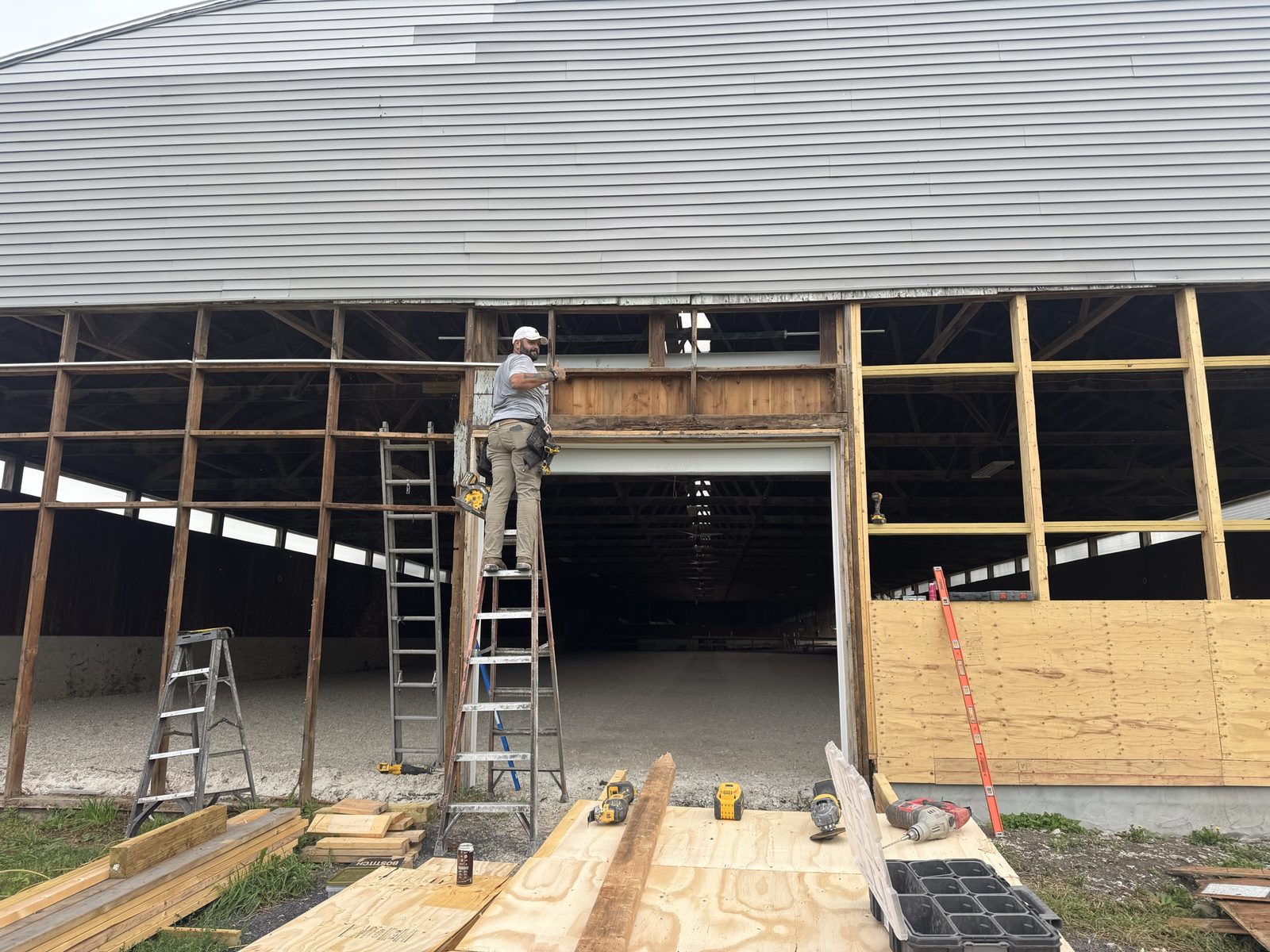 Full view of a barn gable end under restoration with a crew member on a ladder repairing framing above the door opening