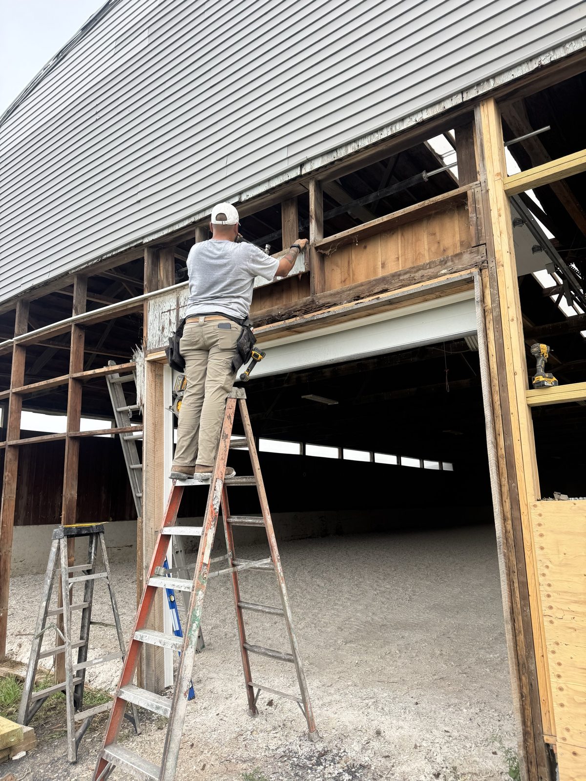 Crew member on a ladder removing rotted fascia board above a barn gable doorway during restoration