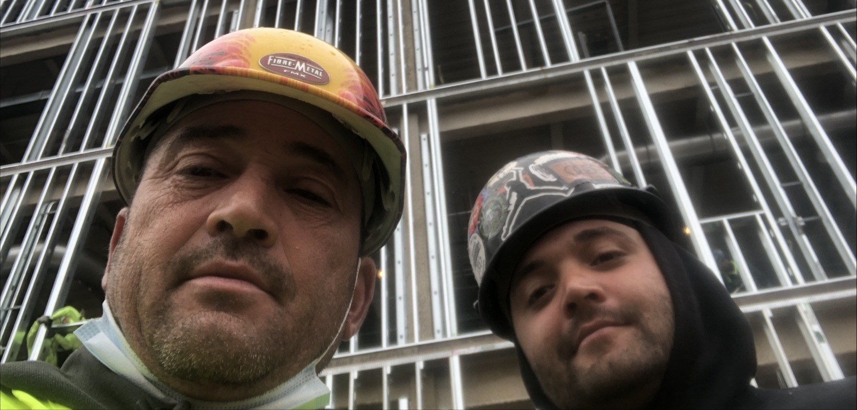 Two crew members taking a selfie in front of a steel-framed commercial building under construction